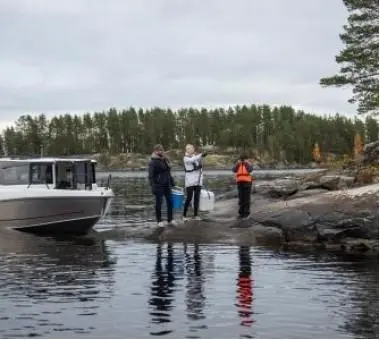 Three people stand on a rocky shoreline by a lake, alongside a boat, preparing fishing gear in a scenic natural setting.